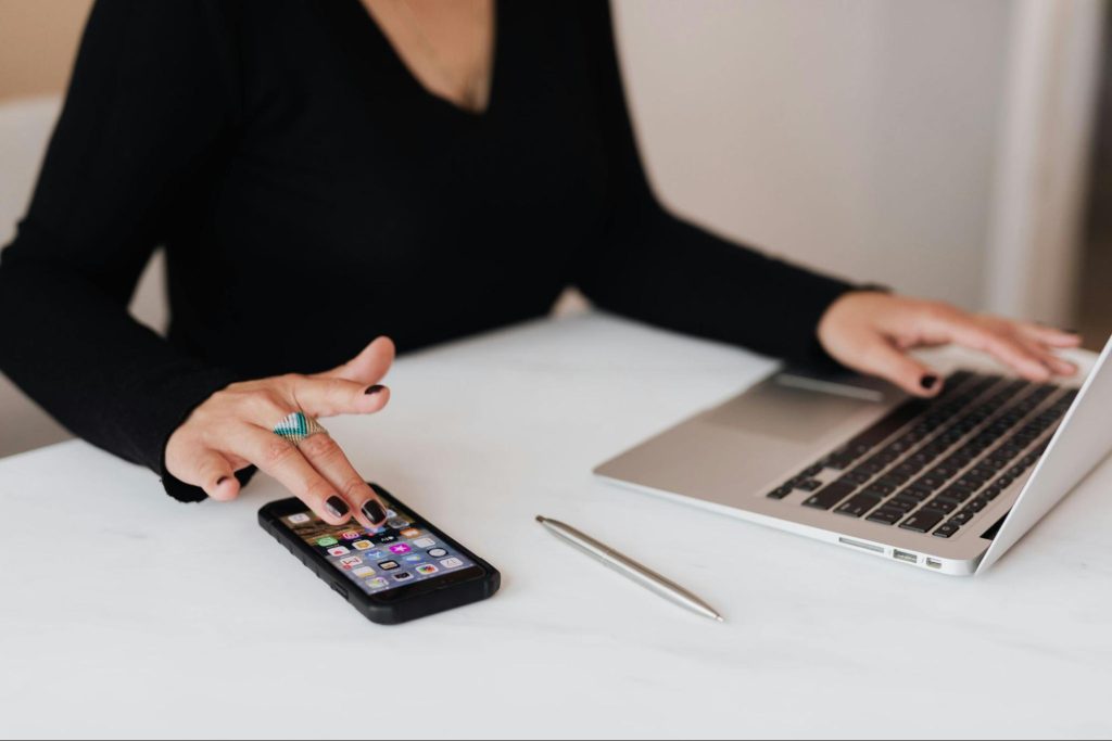 A woman wearing black long sleeves is hovering her left hand on a laptop while her right hand is tapping on a mobile phone.