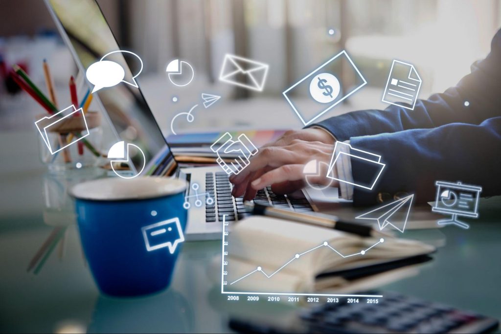 A photo of a man’s hands typing on a laptop keyboard with icons surrounding it, such as a folder, pie graph, link graph, cash, mail, etc.